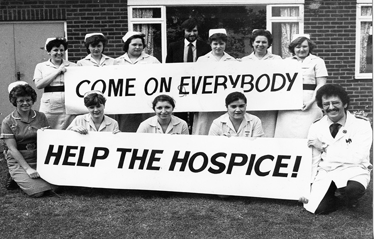 Nurses and doctors holding up a sign that says 'Come on everybody, help the hospice'