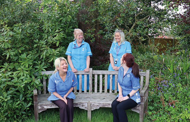 St Helena Virtual Ward staff and Bluebird Care staff sitting on a bench enjoying light conversation. They are smiling and laughing.